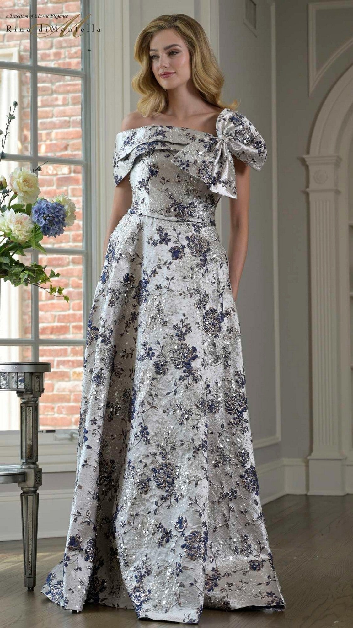 A woman in a navy Rina Di Montella RD3425 long formal dress with an off-shoulder neckline, floral brocade pattern, bow detail, and elegant A-line skirt stands indoors by a window and floral arrangement.